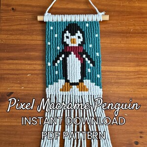 Könnte beinhalten: Ein Makramee-Wandbehang mit einem verpixelten Pinguin-Motiv in Schwarz, Weiß, Blaugrün und Orange, der an einem Holzstab mit einer weißen Kordel hängt. Der Text Pixel Macrame Penguin INSTANT DOWNLOAD PDF PATTERN ist über das Bild gelegt.