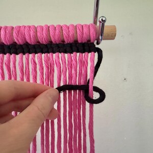 Puede incluir: Primer plano de un tapiz de pared tejido en rosa y negro. La secci&oacute;n superior est&aacute; hecha de hilo rosa bien enrollado, con una secci&oacute;n negra debajo. Largos mechones de hilo rosa cuelgan. Se muestra una mano trabajando con el hilo negro.