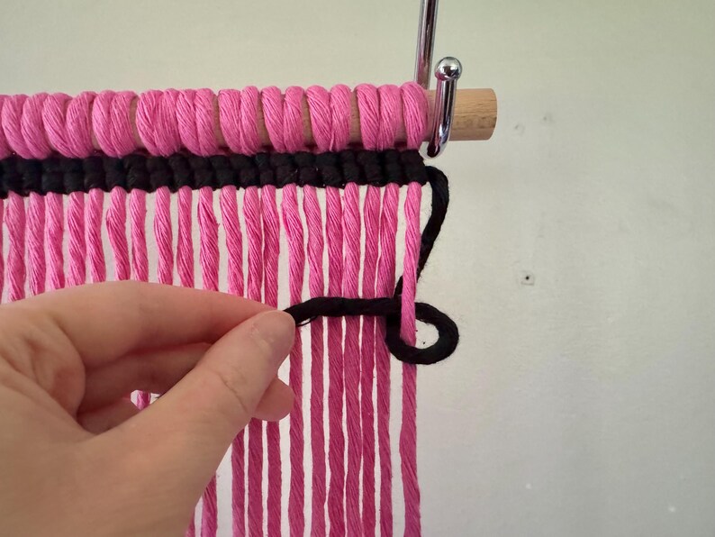 Puede incluir: Primer plano de un tapiz de pared tejido en rosa y negro. La secci&oacute;n superior est&aacute; hecha de hilo rosa bien enrollado, con una secci&oacute;n negra debajo. Largos mechones de hilo rosa cuelgan. Se muestra una mano trabajando con el hilo negro.