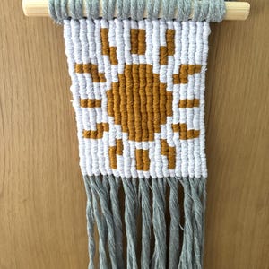 May include: A handmade macrame wall hanging featuring a sun design in mustard yellow and white. The wall art is suspended from a wooden dowel and has gray fringe at the bottom. The piece is a decorative textile.