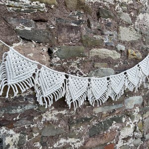 Op de afbeelding: Een witte macramé banner met een reeks driehoekige vormen en franjes hangt tegen een verweerde stenen muur. De banner is gemaakt van geknoopt katoenen koord en wordt opgehangen door een dun wit koord.