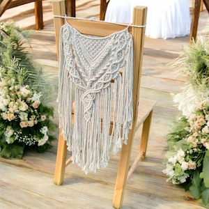Può includere: Una decorazione per lo schienale della sedia in macramé bianco con un motivo geometrico e frange. La decorazione è appesa a una sedia di legno.