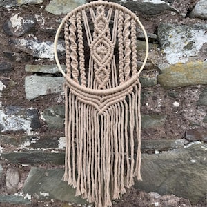 May include: A beige macrame wall hanging with a circular wooden frame. The design features intricate knotting patterns, including a central diamond shape and vertical strands that cascade downwards. The wall decor is suspended by a matching cord.
