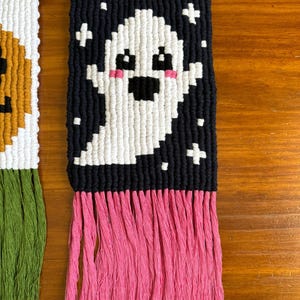 Puede incluir: Un tapiz de macram&eacute; hecho a mano con un dise&ntilde;o de fantasma blanco sobre un fondo negro con acentos de cruz blancos. El fantasma tiene mejillas rosadas y rasgos negros. Tiene una franja rosa en la parte inferior y est&aacute; unido a una varilla de madera.