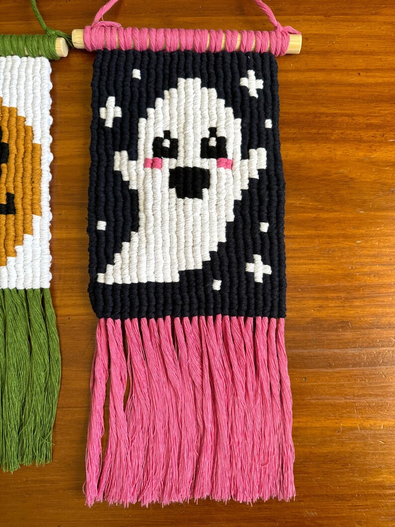 Puede incluir: Un tapiz de macram&eacute; hecho a mano con un dise&ntilde;o de fantasma blanco sobre un fondo negro con acentos de cruz blancos. El fantasma tiene mejillas rosadas y rasgos negros. Tiene una franja rosa en la parte inferior y est&aacute; unido a una varilla de madera.