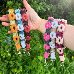 May include: A selection of colourful macrame wrist straps, each handmade with crocheted flower embellishments. The straps display a range of hues, such as orange, green, white, pink, and burgundy, and are finished with silver clasps.