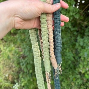 May include: Close-up of three macrame straps in shades of green, beige, and teal. Each strap has a silver clasp. The straps are held against a blurred green background of foliage.