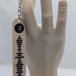 Könnte beinhalten: Ein beigefarbener Schlüsselanhänger mit einem schwarzen Spotify-Code-Design. Der rechteckige Schlüsselanhänger hat abgerundete Kanten und ist an einer silbernen Kette befestigt. Das Spotify-Logo befindet sich oben. Der Hintergrund ist hellbeige.