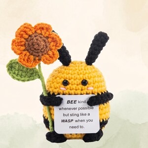 May include: A crocheted bee figure, yellow with black stripes and antennae, holding a sunflower and a sign. The sunflower is orange and brown, with a green stem and leaf. The sign reads "BEE kind whenever possible but sting like a WASP when you need to."