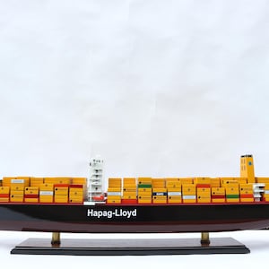 May include: A model of a cargo ship with the text "Hapag-Lloyd" on the side. The ship is black with a red hull and yellow containers stacked on top. The model sits on a dark wooden base.