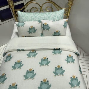 May include: Miniature bed with a gold-colored metal frame. The bedding features a white background with a pattern of light blue frogs wearing gold crowns. Two small pillows with frog designs and a patterned bolster pillow complete the set.