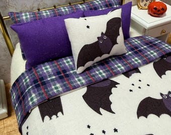 May include: A miniature bed set featuring a white blanket with a repeating black bat and star pattern, a purple and green plaid runner, two solid purple rectangular pillows, and one small square pillow with a central black bat graphic.