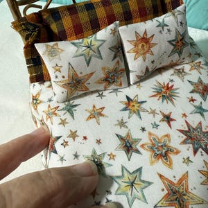 May include: Miniature bed with a gold-colored metal frame. The bedding features a white fabric with colorful star patterns in shades of blue, orange, and green. Two small pillows and a patterned headboard complete the cozy scene.