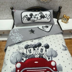 Puede incluir: Un juego de cama en miniatura que incluye una almohada gris con el texto 1928 y una ilustración de Mickey y Minnie Mouse. La manta gris y blanca muestra un patrón de estrellas, puntos y Mickey y Minnie conduciendo un coche rojo.