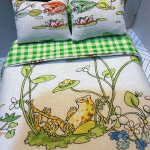 May include: Miniature doll bed with frog-themed bedding. The bed features two small pillows with frog and lily pad designs. The bedspread has a green and white gingham pattern, and the bottom sheet has a frog and lily pad illustration.