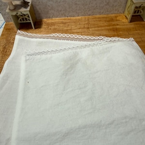 May include: A folded white linen cloth with delicate lace trim along the top edge. The fabric appears soft and slightly textured. The lace trim is a light brown color. The cloth is likely a decorative or functional textile.