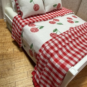 May include: Miniature white bed with a cherry-themed bedding set. The set includes two pillows, a comforter, and a bedspread. The comforter and pillows feature a white background with red cherries and green stems. The bedspread is red and white checkered.