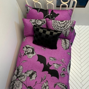 May include: Miniature bed with a white metal frame and purple bedding. The bedding features a floral and bat pattern in black and white. A small black checkered pillow rests on top of the bed.