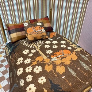 May include: A miniature bed with a brown fox-themed bedding set and pillows. The bedspread features a repeating pattern of white flowers and sleeping foxes. The pillows have plaid and fox designs. The bed frame is gold-colored.