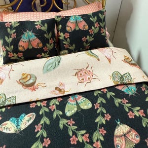 Puede incluir: Juego de cama en miniatura con marco de metal dorado, con almohadas y edredón con estampados de insectos y flores. Las almohadas son negras con polillas coloridas y flores rosas. El edredón tiene un fondo claro con varios insectos y mariposas.