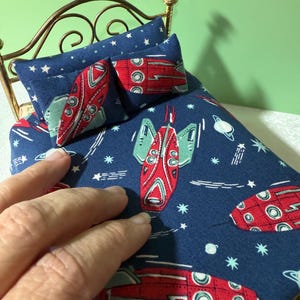 Puede incluir: Cama en miniatura con marco de latón y ropa de cama con temática espacial. La tela azul marino presenta cohetes rojos, estrellas blancas y planetas. Dos almohadas a juego descansan sobre la cama. Se ve una mano tocando la ropa de cama.