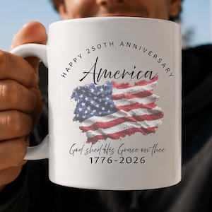 May include: A white ceramic mug featuring a graphic of the American flag with the text Happy 250th Anniversary America, God shed His Grace on thee, and the years 1776-2026 printed in black ink on the front surface of the cylindrical vessel.
