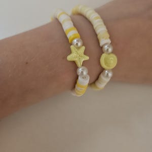 May include: Two yellow and white beaded bracelets. One bracelet features a yellow star charm and the other has a yellow seashell charm. Both bracelets have pearl accents and are worn on a wrist.