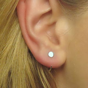 May include: A close-up of a person's ear with a small, round, silver earring. The earring is a simple stud with a smooth, shiny surface.