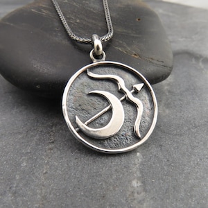 May include: A silver pendant necklace featuring a circular pendant with a crescent moon and bow and arrow design. The pendant hangs from a silver chain. The pendant is displayed on a dark gray stone surface.