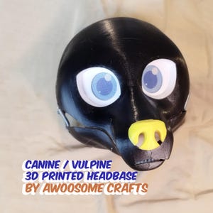Puede incluir: Una base de cabeza canina o vulpina negra impresa en 3D con ojos grandes de color azul claro y una nariz amarilla. El texto "CANINE / VULPINE 3D PRINTED HEADBASE BY AWOOSOME CRAFTS" está impreso en la parte inferior de la imagen.