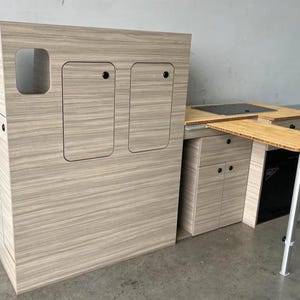 Op de afbeelding: Een licht houten kast met twee deuren en een klein raam. De kast is bevestigd aan een aanrechtblad met een neerklappende tafel. De tafel is gemaakt van licht hout en heeft een witte metalen steun.