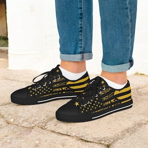 Puede incluir: Zapatillas de lona negras con un diseño de bandera dorada y negra. Los zapatos de corte bajo tienen cierre de cordones y suela blanca. El diseño incluye estrellas y rayas, creando un tema patriótico.