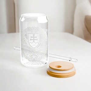 Op de afbeelding: Een helder glazen drinkglas in de vorm van een blikje met een geëtst ontwerp, een glazen rietje en een houten deksel. Het ontwerp op het glas heeft een wapen met een kruis en andere sierlijke details. De deksel is van licht hout.