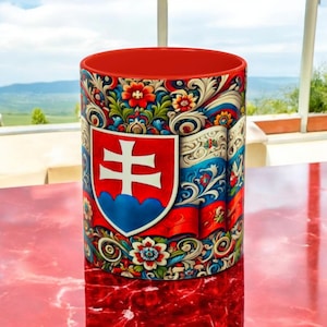 May include: A red and white coffee mug with a floral design and the Slovakian coat of arms. The mug is on a red surface.