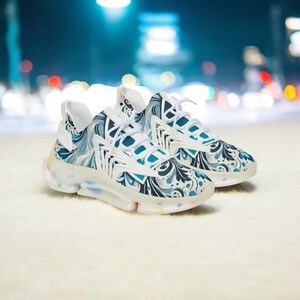 Puede incluir: Un par de zapatillas deportivas con un estampado floral blanco y azul. Las zapatillas tienen cordones blancos y una suela translúcida con detalles coloridos. El fondo está borroso, sugiriendo un entorno al aire libre.