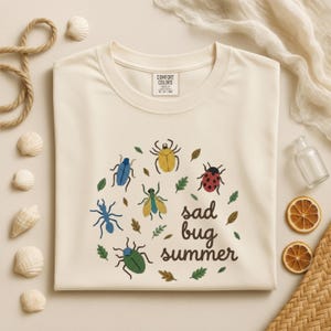 Può includere: T-shirt color crema con un design circolare di insetti e foglie di cartoni animati colorati. Le parole "sad bug summer" sono stampate in un carattere stravagante. La maglietta è realizzata in materiale morbido e ha uno scollo tondo.