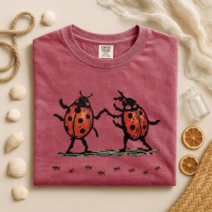 Pode incluir: Uma camiseta Comfort Colors bordô com um gráfico de duas joaninhas dançando com bolinhas laranja e pretas. A camisa tem uma pequena etiqueta Comfort Colors. Pequenas ilustrações de formigas na parte inferior.