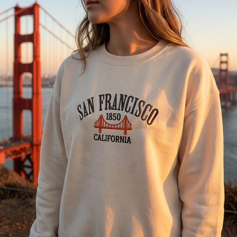 May include: A cream-colored crewneck sweatshirt featuring an embroidered design on the chest. The text reads SAN FRANCISCO 1850 CALIFORNIA in black lettering, accompanied by an orange embroidered graphic of the Golden Gate Bridge.
