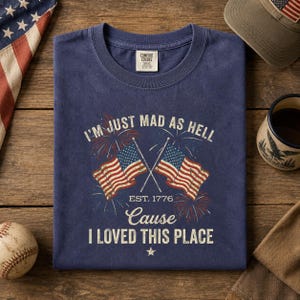 Może przedstawiać: Granatowa koszulka z napisem "I'M JUST MAD AS HELL CAUSE I LOVED THIS PLACE" i dwiema amerykańskimi flagami. Koszulka zawiera również napis "EST. 1776". Widoczna jest również piłka do baseballu, czapka z amerykańską flagą i kubek.