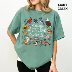 Könnte beinhalten: Ein hellgrünes Kurzarm-T-Shirt mit einem Grafikdruck von verschiedenen Vögeln, einem Vogelhaus, einem Fernglas, Blumen und einem Nest. Der Text "easily distracted by birds" ist mittig in weißer Schreibschrift aufgedruckt. Rundhalsausschnitt.