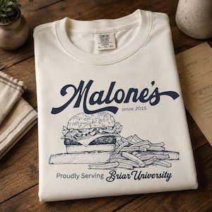 May include: Cream-colored t-shirt with a navy blue graphic of a burger and fries, and the text "Malone's since 2015". Below the image is the text "Proudly Serving Briar University".