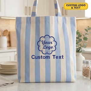 May include: A vertical striped tote bag in light blue and off-white fabric. The center features an embroidered dark blue scalloped circular emblem containing the text Your Logo and the words Custom Text printed below the emblem.