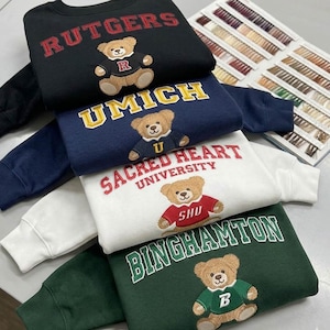 May include: A stack of sweatshirts in black, navy, white, and green, each embroidered with a university name and a teddy bear mascot. The sweatshirts feature the names "RUTGERS", "UMICH", "SACRED HEART UNIVERSITY", and "BINGHAMTON".