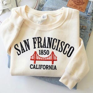 May include: A cream-colored crewneck sweatshirt featuring black embroidered text that reads San Francisco 1850 California. Below the text is an embroidered red graphic of the Golden Gate Bridge. The sweatshirt is folded over blue denim jeans.