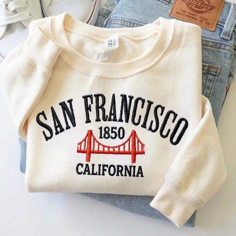 May include: A cream-colored crewneck sweatshirt featuring black embroidered text that reads San Francisco 1850 California. Below the text is an embroidered red graphic of the Golden Gate Bridge. The sweatshirt is folded over blue denim jeans.