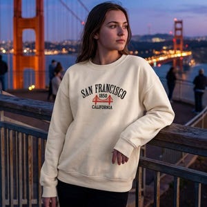 May include: A cream-colored crewneck sweatshirt featuring black text that reads SAN FRANCISCO CALIFORNIA with a red graphic of the Golden Gate Bridge and the year 1850 printed in the center of the chest on the front of the garment.