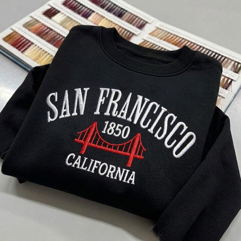 May include: A black crewneck sweatshirt featuring white embroidered text that reads SAN FRANCISCO and CALIFORNIA, with a red embroidered graphic of the Golden Gate Bridge and the year 1850 positioned between the two lines of text.