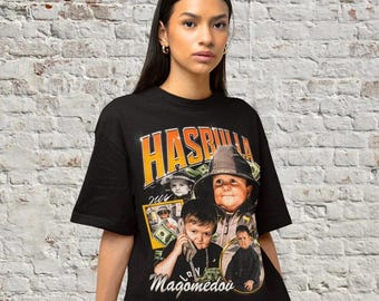 May include: Black t-shirt with a graphic print of a young man wearing a hat and surrounded by money. The text "HASBULLA" and "Magomedov" are printed on the shirt.
