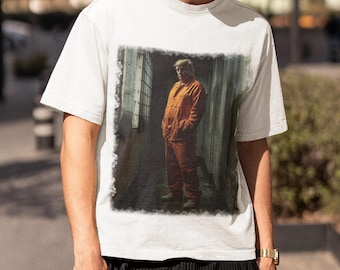 May include: A white t-shirt with a faded image of a man in an orange jumpsuit standing in a hallway. The image is surrounded by a dark border.