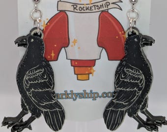 May include: A pair of black raven earrings with detailed feather designs. The earrings hang from silver hooks. The background features a white card with a rocket ship graphic and the text "Sparkly Rocketship".